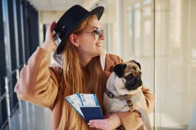Young woman traveling with her dog tips her hat as she holds her pug and passports in her arms.